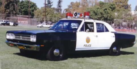 1991 Ford LTD Crown Victoria - Restored black-and-white Plymouth Belvedere police sedan parked on a grass field, wearing 'POLICE' door lettering with a gold city shield d…