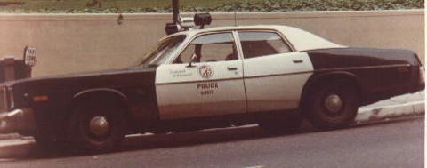 1991 Ford LTD Crown Victoria - Vintage side-view photo of a black-and-white 1970s Mopar four-door police sedan marked LAPD, parked at a curb along a concrete retaining wa…