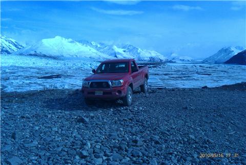2009 Toyota Tacoma - A red 2009 Toyota Tacoma pickup truck is parked on a rocky surface with a vast glacial landscape and snow-covered mountains behind it.