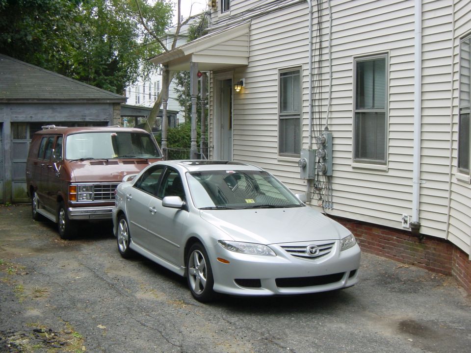 Front three-quarter view of a silver Mazda 6 sedan in a driveway, with a brown van parked behind it