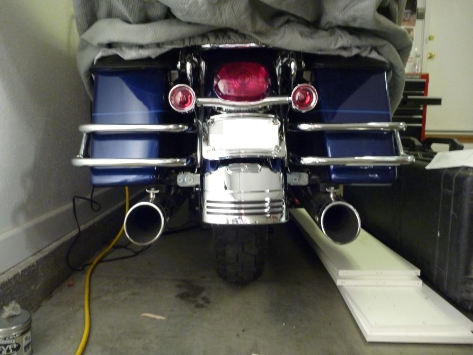 Rear view of a blue motorcycle with saddlebags, chrome trim, and dual exhaust pipes. A grey cover is draped over the front
