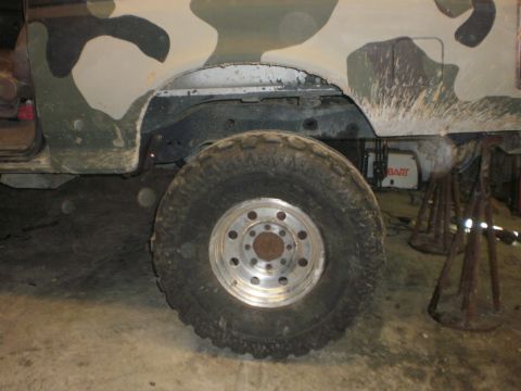 1995 Ford Bronco - Close-up of a vehicle's rear wheel and fender, showing camouflage paint and some mechanical components underneath.
