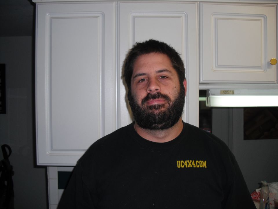 Man with beard wearing black t-shirt with yellow text UC4X4.COM. White kitchen cabinets in background