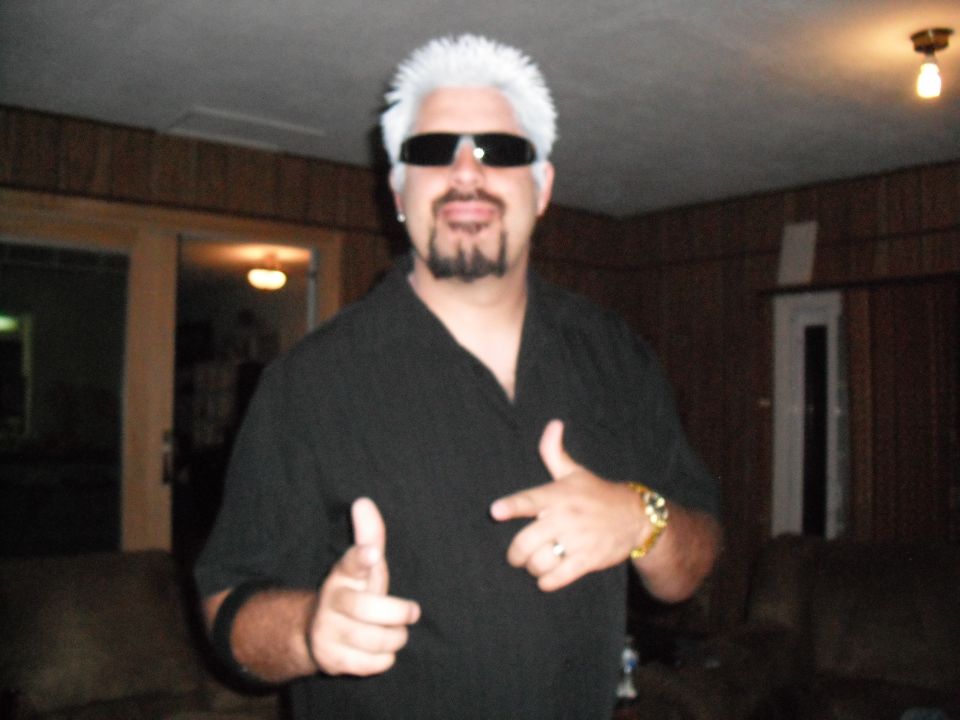 Man in black shirt, sunglasses, and white spiky hair points forward with both hands