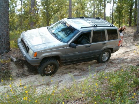1991 Ford Bronco - Silver Jeep Grand Cherokee navigates a sandy trail in a wooded area.