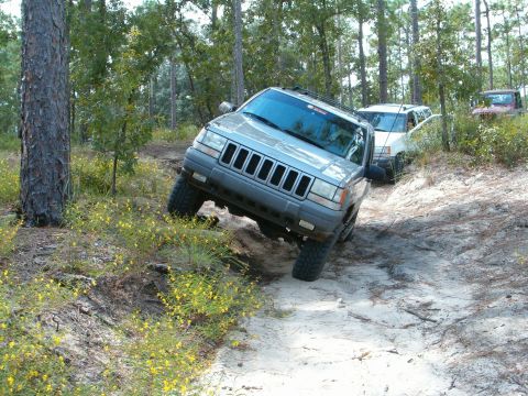 1991 Ford Bronco - A Jeep Grand Cherokee tackles a challenging off-road course, with other vehicles following on the sandy path.