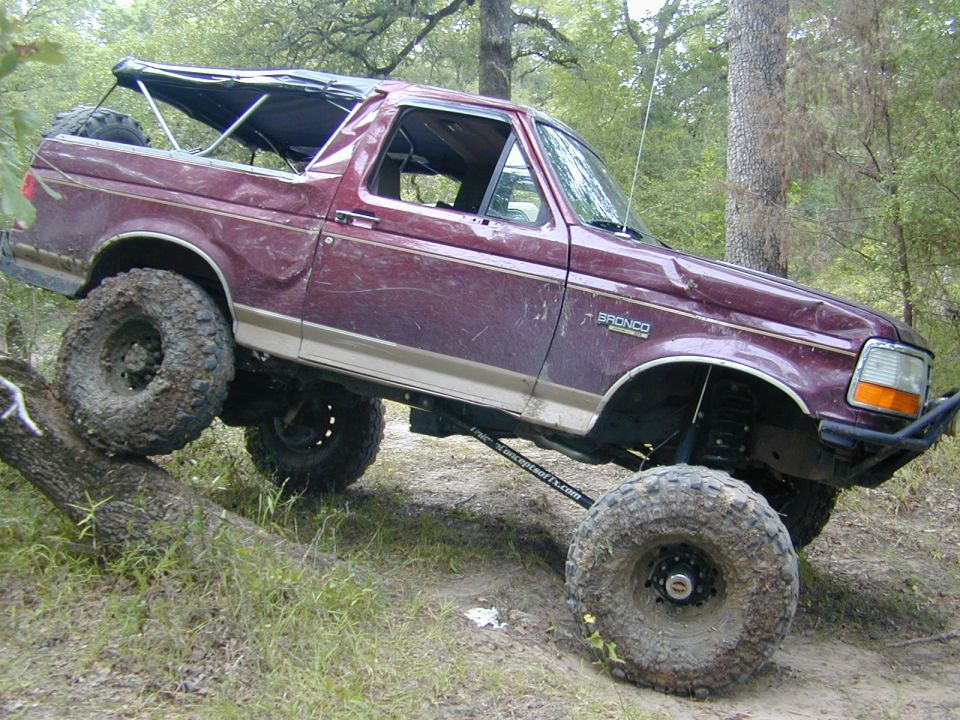 Maroon lifted Ford Bronco on big muddy tires climbing a log in the woods