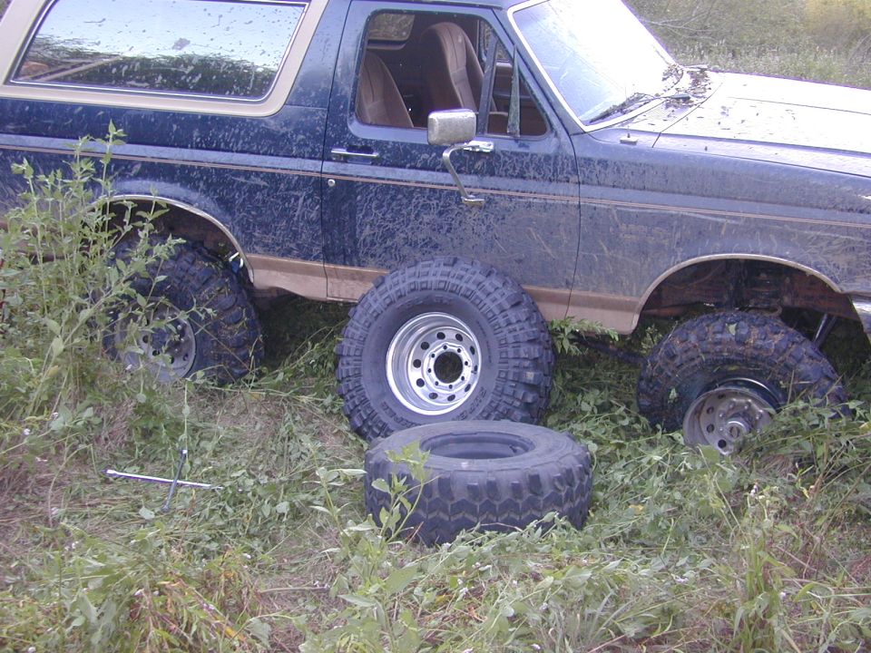 Muddy dark blue Ford Bronco in tall weeds with spare mud-terrain tires and wheels leaning against its passenger side