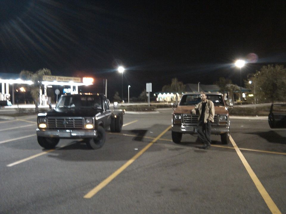 Black Ford dually flatbed and brown square-body pickup parked at night with a man leaning on the fender