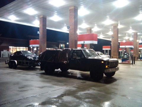 1984 Ford Bronco II - A dark-colored Ford F-Series truck is seen towing a car on a trailer at a gas station at night.