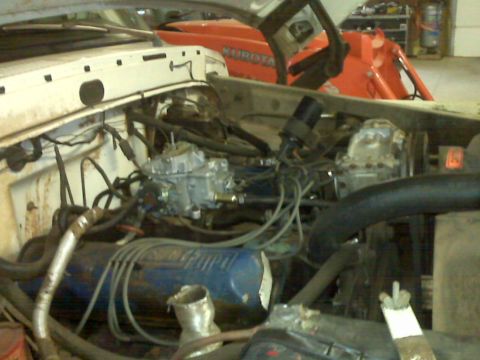 1973 Ford F-100 - Engine bay of a white truck with a blue valve cover and dual carburetors. Orange tractor in background.