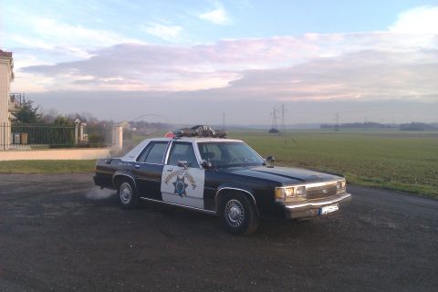 1991 Ford LTD Crown Victoria - Black-and-white Ford LTD Crown Victoria patrol car parked on a gravel lot beside an open green field at dusk, viewed from the front three-q…