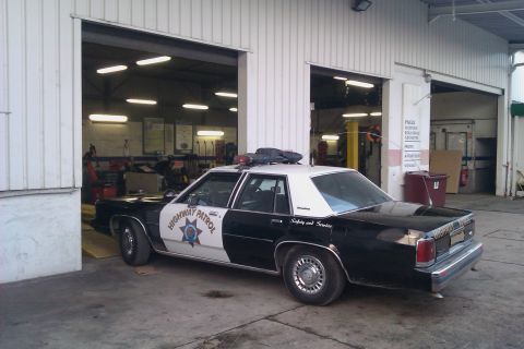 1991 Ford LTD Crown Victoria - Black-and-white Ford LTD Crown Victoria patrol sedan seen from the rear three-quarter outside a tire and service shop with open roller door…