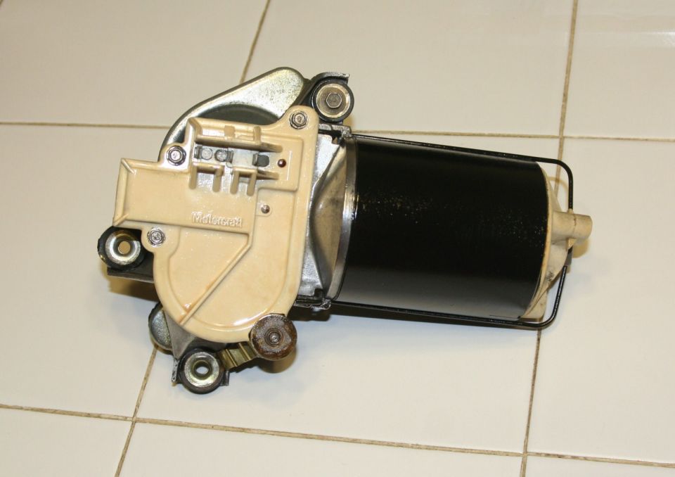 Windshield wiper motor with a black cylinder and beige housing, mounted on a tiled surface