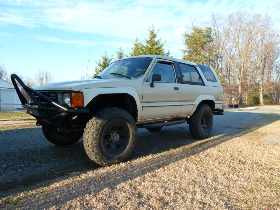Lifted tan Toyota 4Runner on gravel with tube bumper and mud tires, winter trees