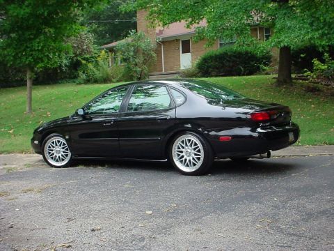 1999 Ford Taurus - A black sedan is parked on the side of the road with a house in the background.