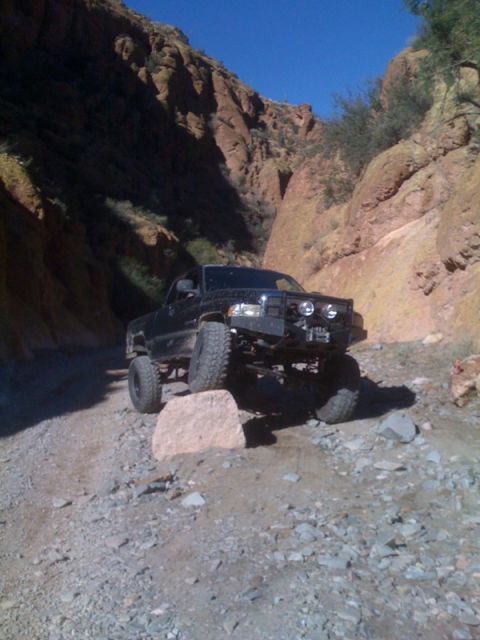1966 Ford Bronco - A lifted black pickup truck navigates a rocky trail through a narrow canyon under a clear blue sky.