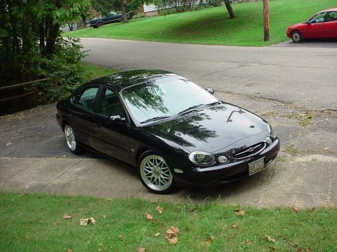 1999 Ford Taurus - A black sedan is parked on a concrete driveway next to a grassy lawn. Another red car is visible in the background.