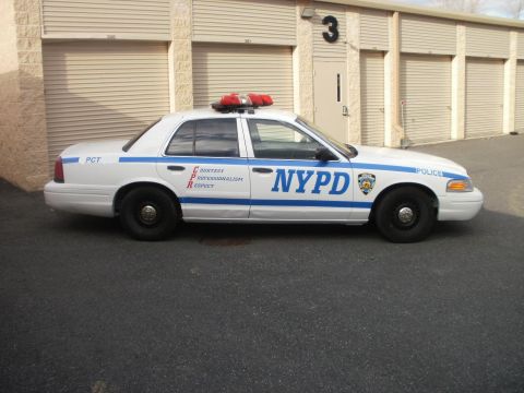 2002 Ford Crown Victoria - A white 2002 Ford Crown Victoria police car with NYPD markings is parked in front of storage units.