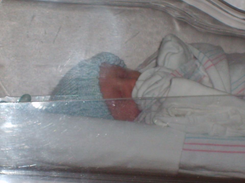 Newborn baby wearing a blue cap and wrapped in a striped blanket inside a clear incubator