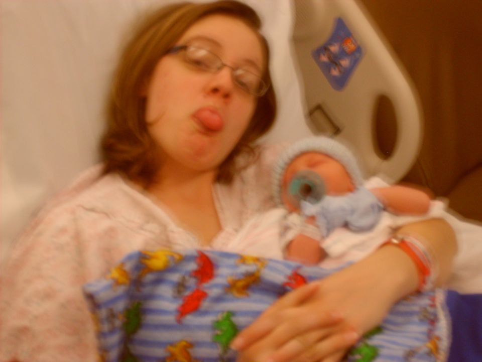 Woman in hospital gown sticking her tongue out while holding a newborn baby with a pacifier