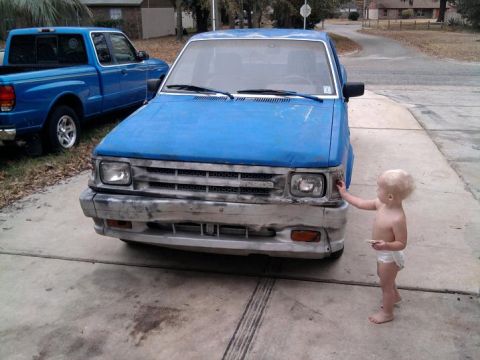 1987 Mazda B2200 - A child interacts with the front of a blue pickup truck, with another truck visible in the background.
