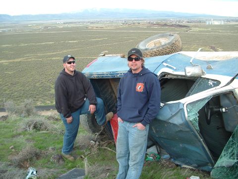 1989 Ford Bronco - Two men pose next to a wrecked, overturned vehicle in a desolate outdoor setting.
