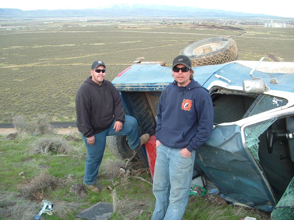 Two guys stand next to a rolled-over blue SUV in a field