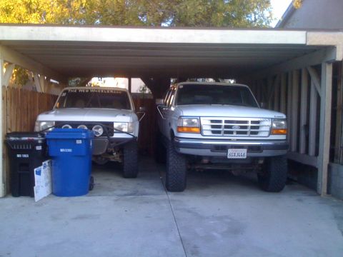 1968 Ford Mustang - Two modified trucks are parked side-by-side in a carport, with trash bins to the left.