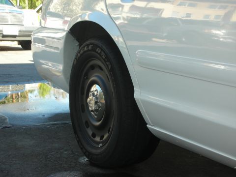 2004 Ford Crown Victoria - A white sedan's rear wheel and lower body panel are visible, with another vehicle in the background.