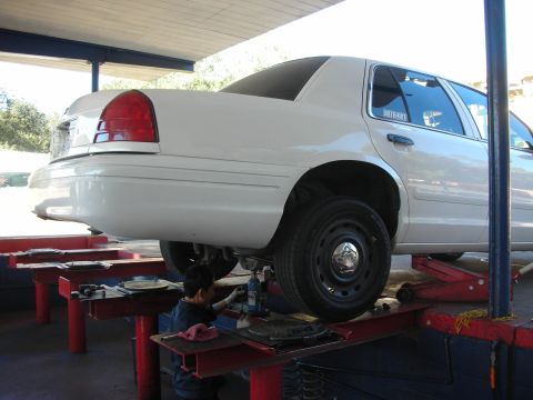 2004 Ford Crown Victoria - A white Ford Crown Victoria sedan raised on a drive-on rack, viewed from the rear three-quarter, with a technician working underneath near…