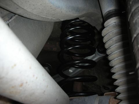 2004 Ford Crown Victoria - Underbody view of a rear coil suspension spring (black, coated) seated between the axle and body, with a ribbed rubber axle boot at right,…
