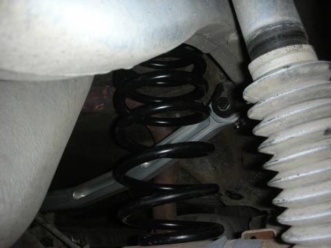2004 Ford Crown Victoria - Close-up under a car showing a black rear coil spring seated on a silver-painted control arm with a hex-head through-bolt at the pivot. A r…