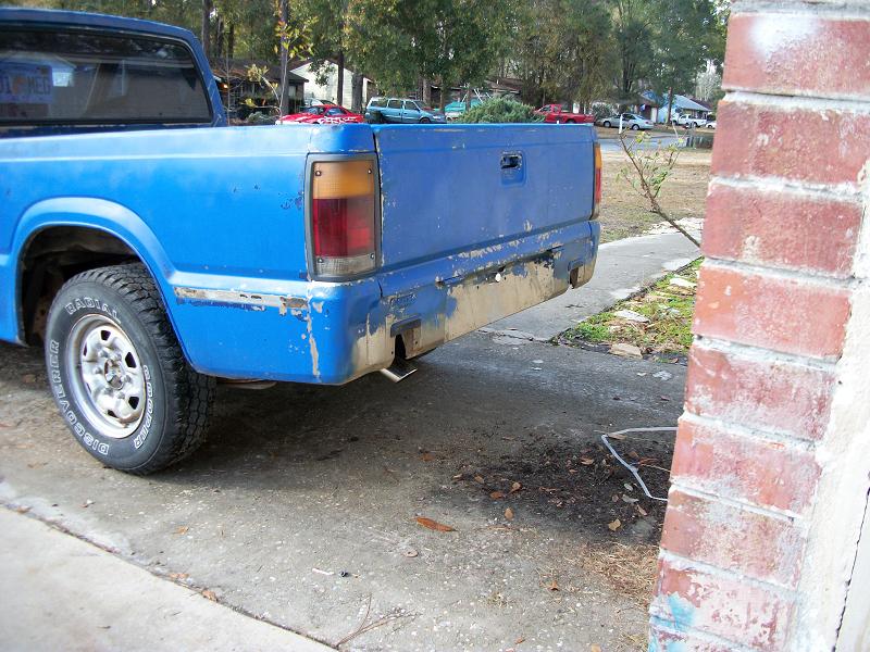 Rear corner of a faded blue Mazda pickup on a driveway near a brick pillar.