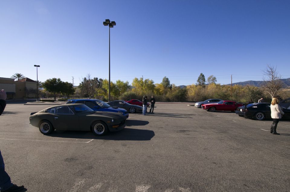Several classic cars parked in a lot, including a dark green coupe in the foreground and a red sports car further back