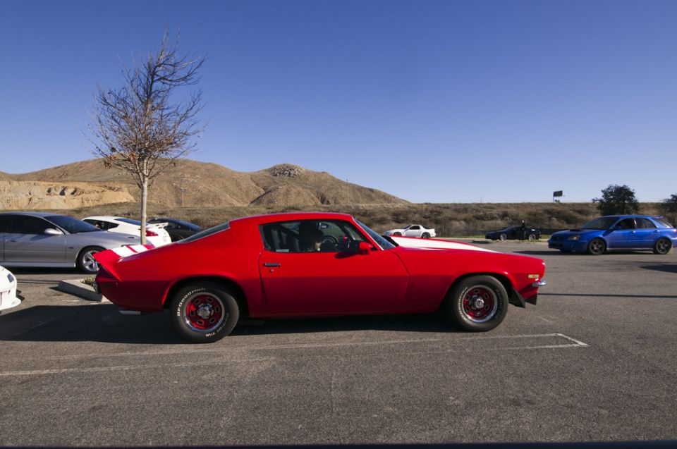 Red 1970s coupe with white hood stripe, side view. Other cars in background