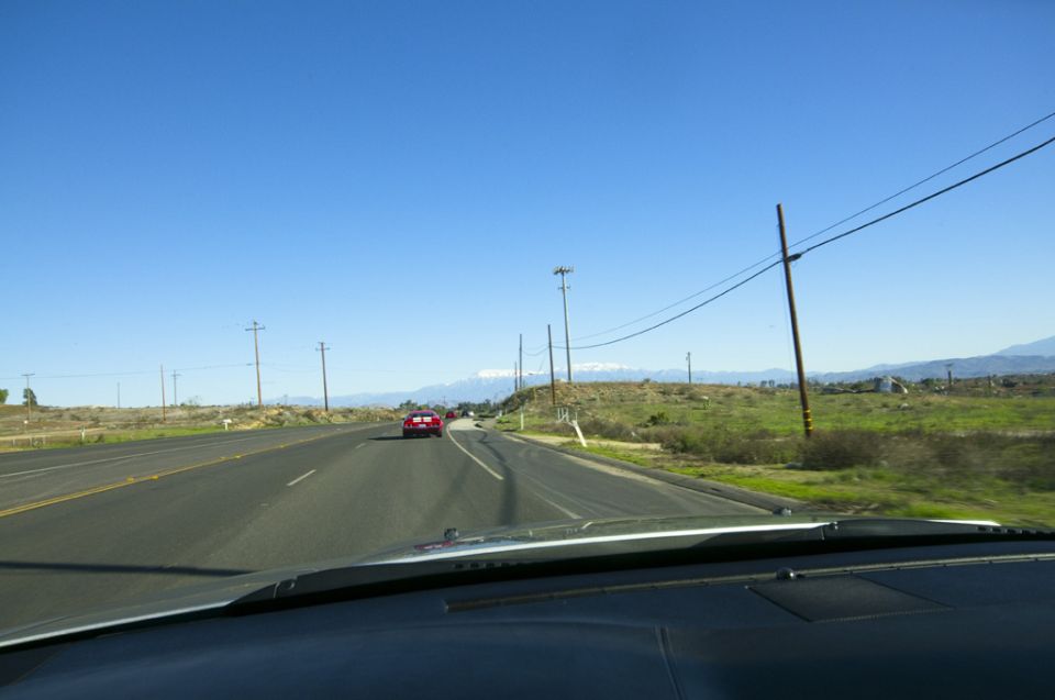 View from inside a car looking out at a highway with a red car in the distance and mountains on the horizon