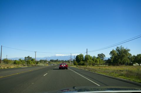 2006 Cadillac STS - A red sports car is seen from behind on a highway, with snow-capped mountains visible in the distance under a clear blue sky.