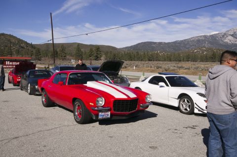2006 Cadillac STS - A line of cars, including a red classic muscle car with white racing stripes, are parked outdoors under a clear sky.