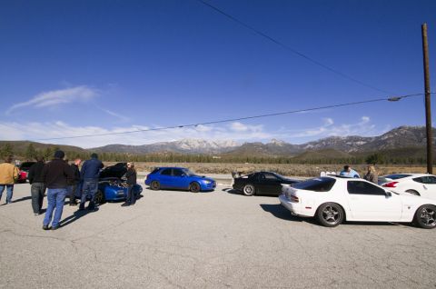2006 Cadillac STS - A group of people stand around several cars in an outdoor lot, with mountains visible in the distance under a clear blue sky.