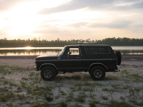 Black vintage Ford Bronco parked on a sandy lakeshore at sunset
