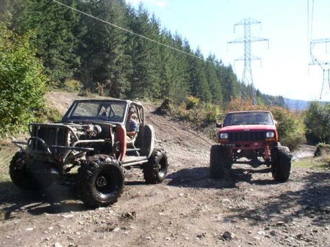 1979 Ford Bronco - Two heavily modified off-road vehicles parked on a dirt trail surrounded by trees.