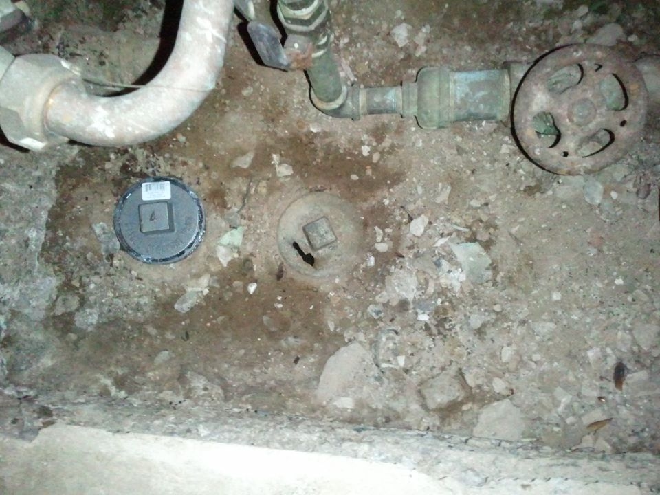 Dirt floor with plumbing pipes, rusted valve wheel and cleanout covers