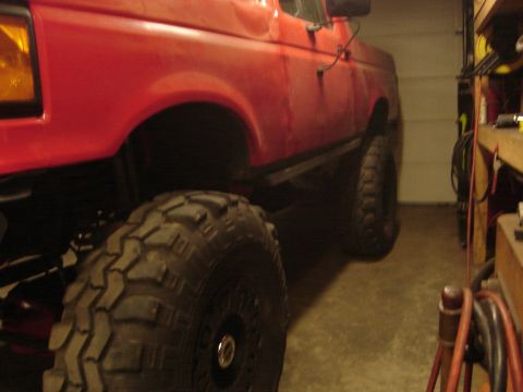 1989 Ford Bronco - Close side view of a lifted red Ford truck in a dim garage, showing tall aggressive mud tires and flat protective rocker armor along the bo…