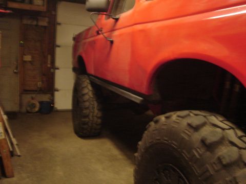 1989 Ford Bronco - Side view of a lifted bright red-orange Ford full-size truck inside a dim garage, riding on very large mud-terrain tires with black angled…