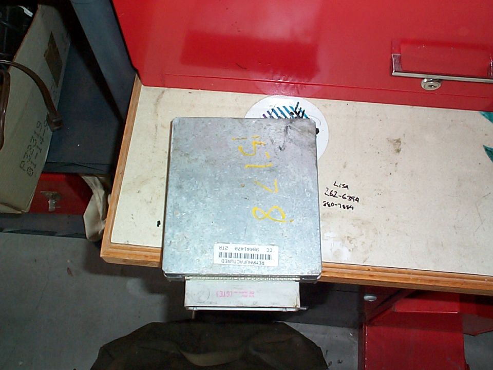 Automotive engine control module with handwritten yellow markings sitting on a wood shelf