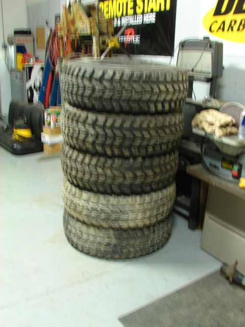 1971 International School Bus - A stack of five large off-road tires sits in a garage, with tools and equipment in the background.