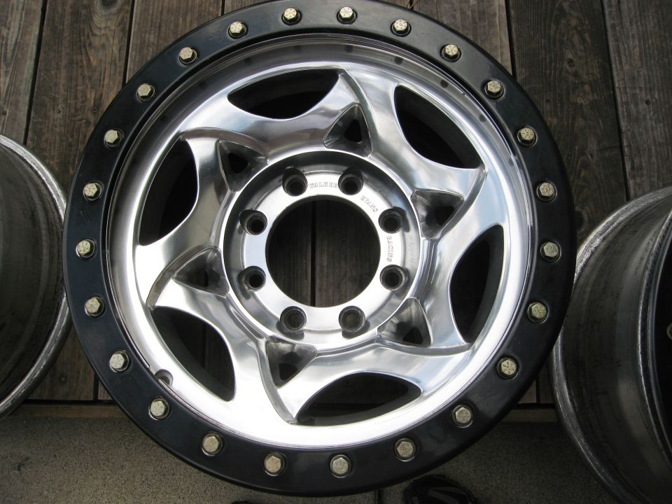 Polished aluminum beadlock wheel with black bolted ring, 8-lug pattern, on wood planks.