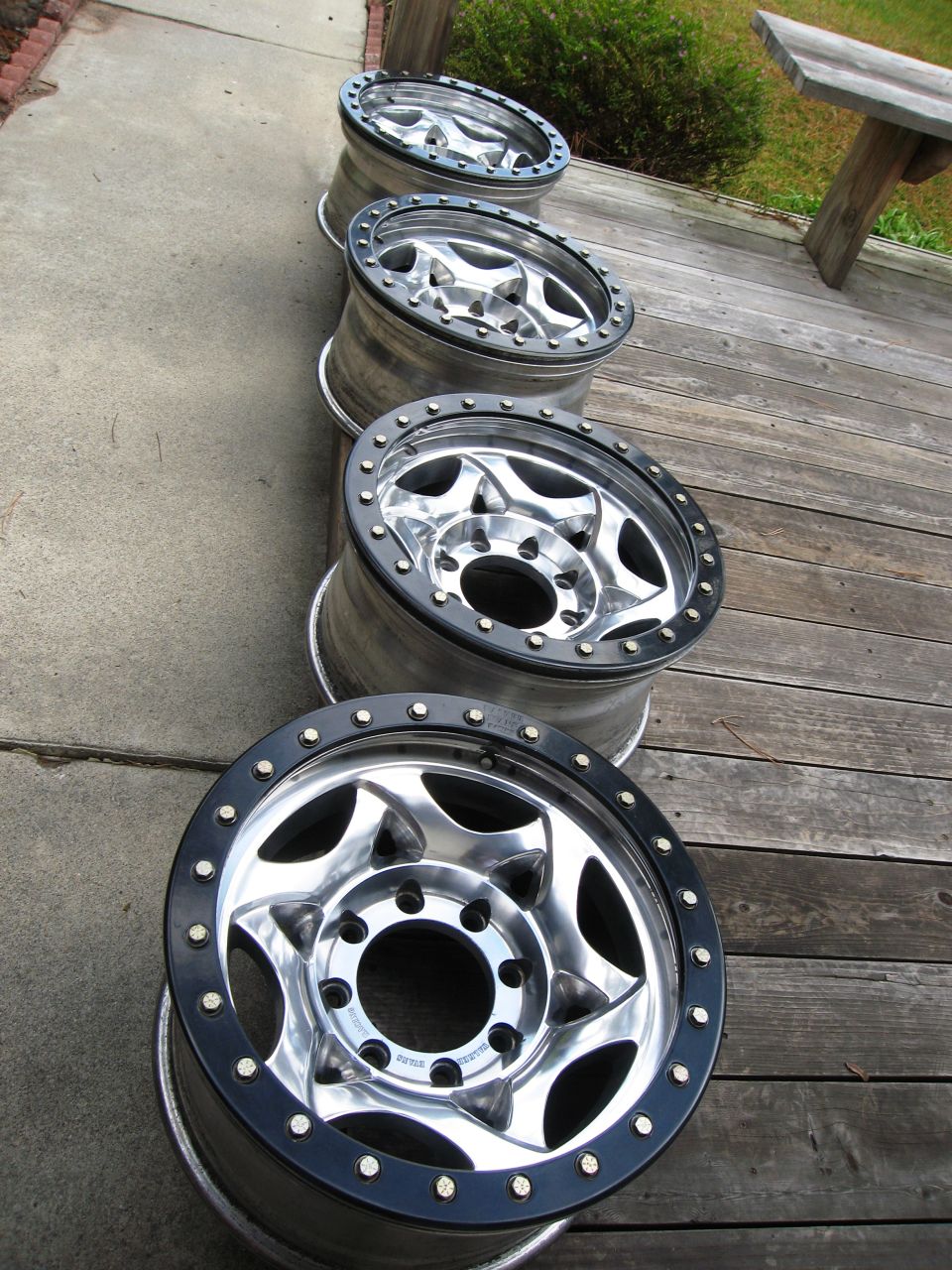 Four polished eight-lug beadlock wheels lined up on a walkway and wood deck