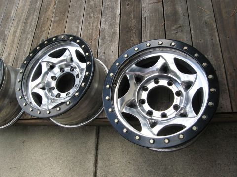 1986 Ford Bronco - Two chrome off-road wheels with black trim are displayed on a weathered wooden surface.